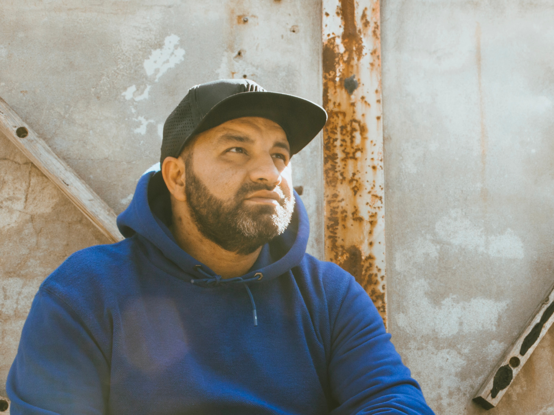 Portret van Alberto Arifi met pet en blauwe hoodie voor een verweerde muur